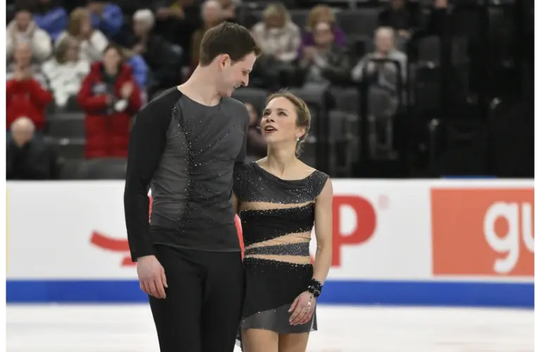 Alisa Efimova and Misha Mitrofanov, a figure skating pair in coordinated dark costumes smiles warmly on the ice. The audience in the background is blurred, creating an intimate, joyful moment getting ready for Team USA and the 2026 Olympics.