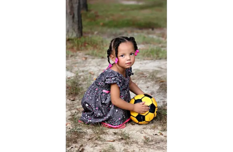 A young girl with braided hair sits on grass, holding a yellow and black soccer ball. She wears a patterned dress with pink accents, conveying a peaceful, thoughtful mood. Showing the hope for National Girls & Women in Sports Day, NGWSD.