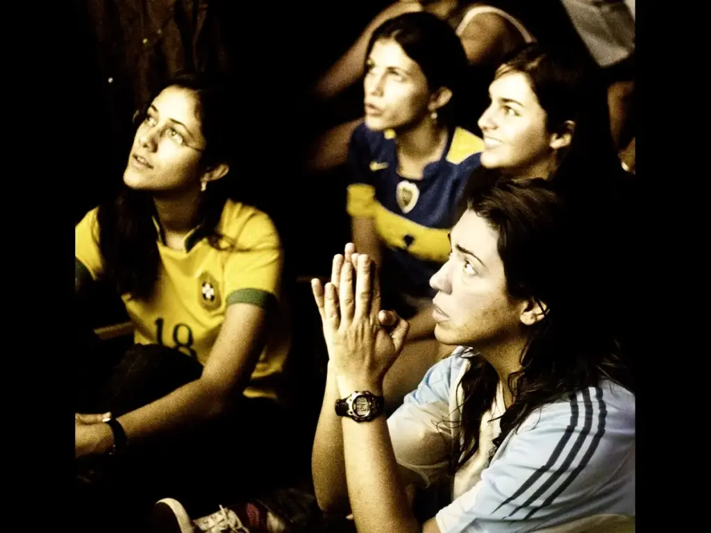 A group of focused women intensely watch a match. They wear different soccer jerseys, displaying anticipation and tension. Overall, the scene is dimly lit.