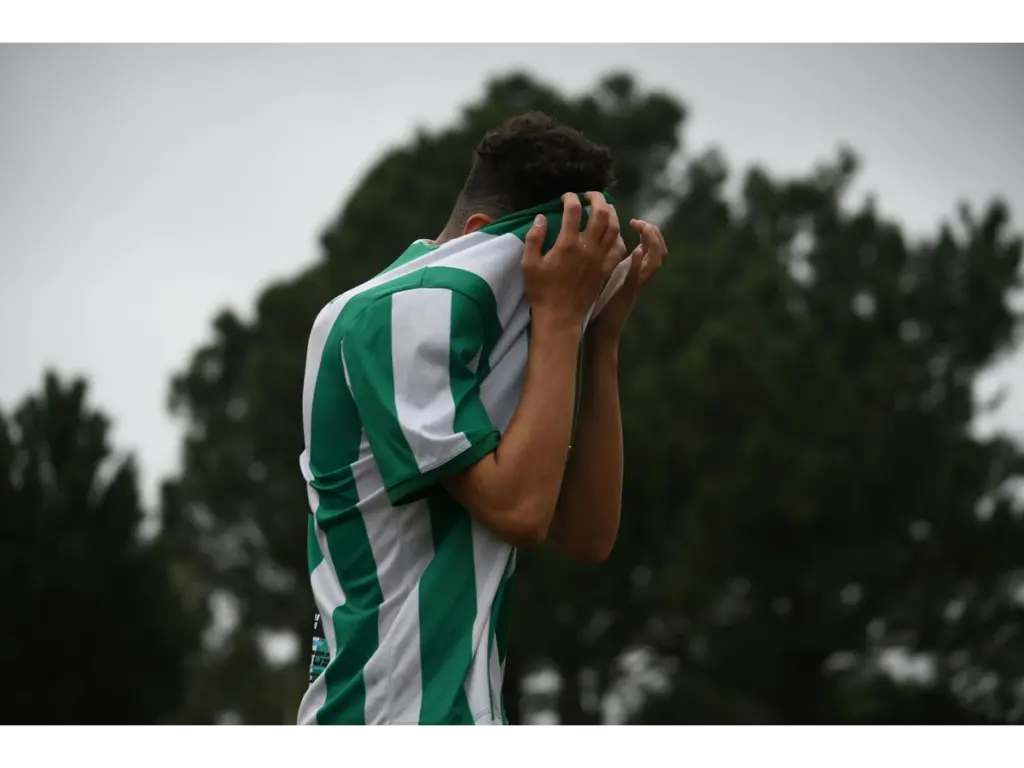 A soccer player in a green and white striped jersey covers their face with the shirt, conveying emotion. Dark trees and overcast sky in the background.