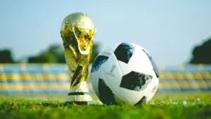 A shiny World Cup gold trophy and an Adidas Telstar soccer ball rest on green grass, with blurred stadium seats in the background under clear blue sky.